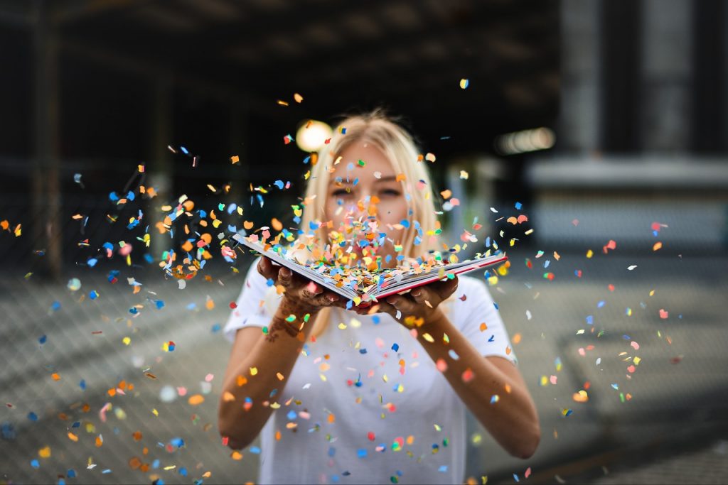 femme blonde en t-shirt balnc qui souffle dans un livre ouvert et fait s'envloer des confettis